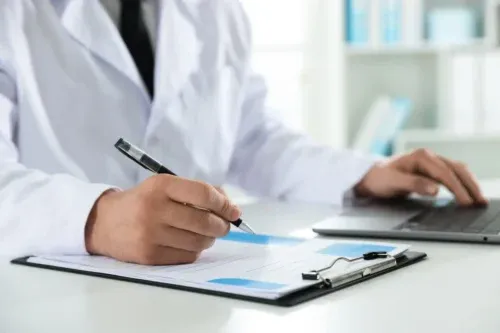 Doctor in medical coat working with clipboard and laptop at white table in clinic, closeup